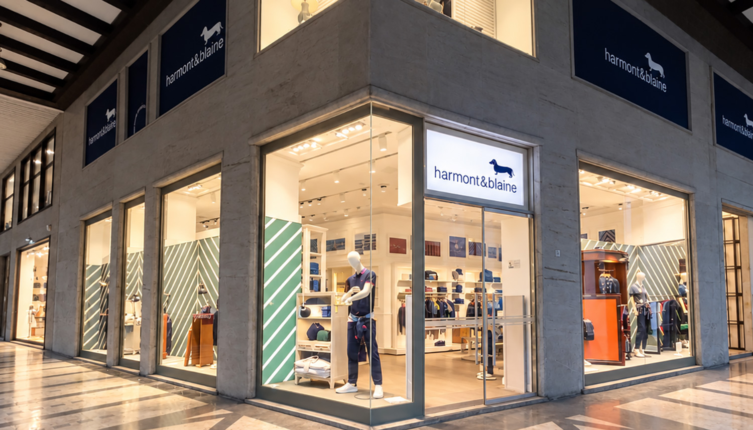 The exterior of a Harmont & Blaine boutique located in a shopping arcade, featuring large corner display windows, warm lighting, and modern interiors with green and white striped walls and displayed mannequins.