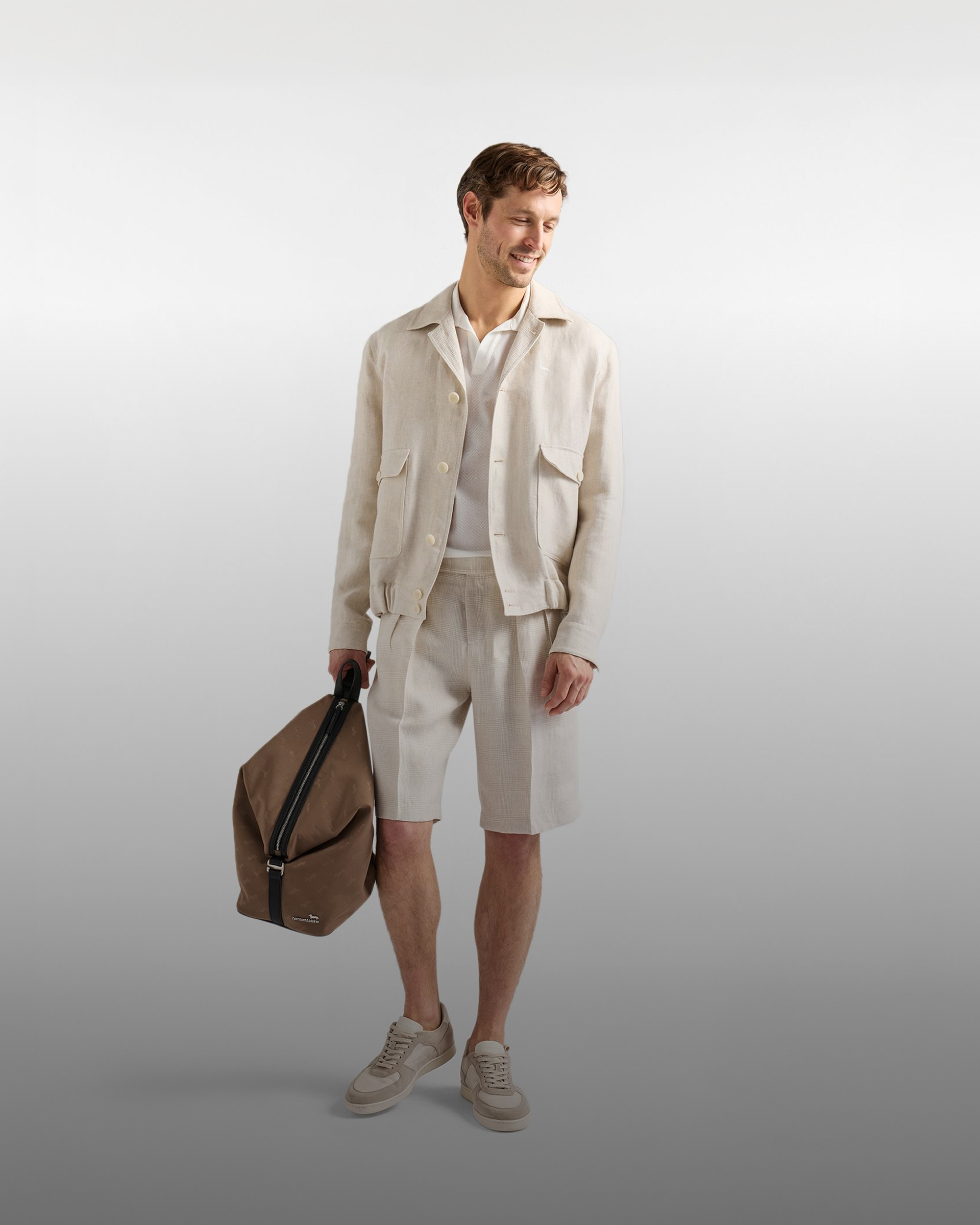 man in a light linen suit with an open jacket, white polo shirt, shorts, and sneakers looks down smiling and holds a brown bag. the background is a gradient gray. at the bottom, the text made in italy and the subtitle the authentic italian spirit appear.