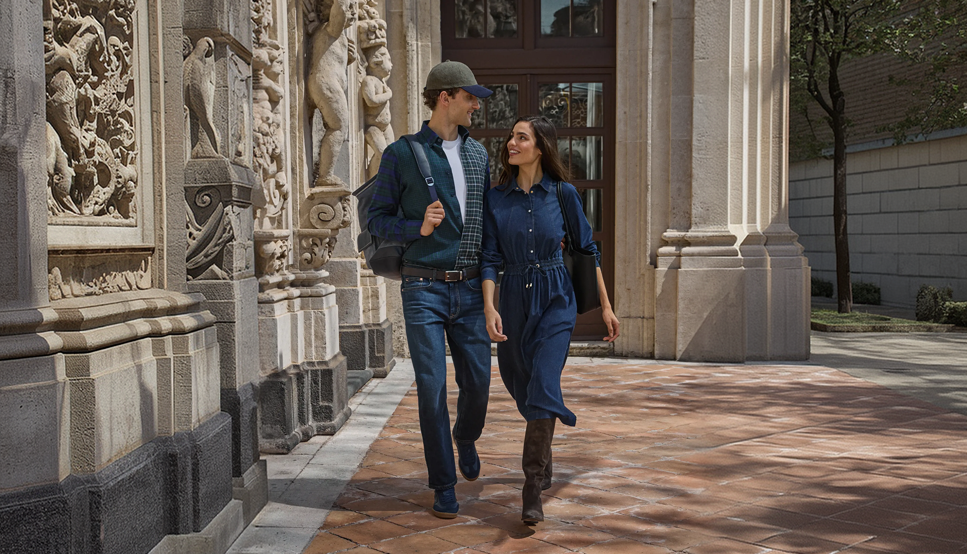 Promotional image for the Fall/Winter 2026 collection featuring a smiling man and woman walking side by side along a historic stone colonnade; the man wears a green and navy tartan cardigan over a white t-shirt, jeans, and an olive green baseball cap, while the woman wears a belted dark blue denim shirt dress, tall brown suede boots, and carries a black shoulder bag.