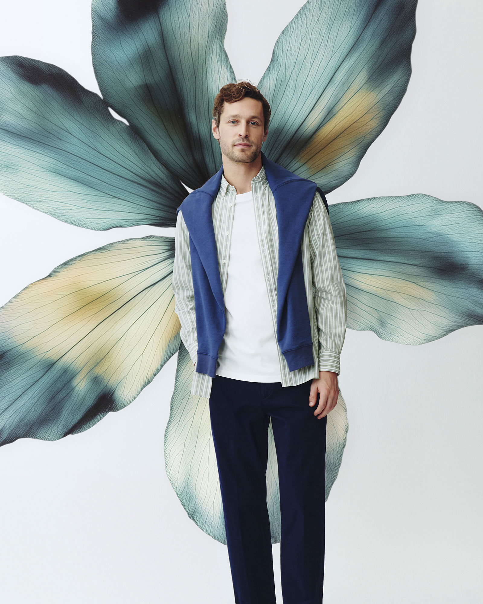 Uomo con camicia a righe verde chiaro, t-shirt bianca, pantaloni blu e maglia blu sulle spalle