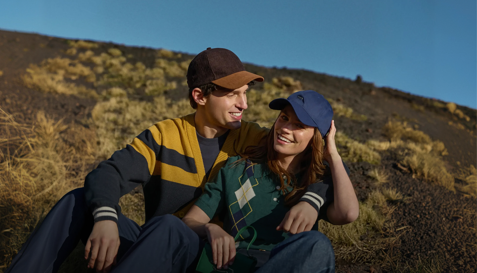This is a campaign image for Harmont & Blaine's Fall/Winter 2026 preview. The image shows a young couple sitting close together outdoors on a grassy, rocky slope under a clear blue sky. The man on the left is wearing a mustard and dark blue color-block ribbed cardigan with contrasting trim on the cuffs, a two-tone brown and light brown cap, and dark trousers. The woman on the right has an arm around the man's shoulders and is wearing a green short-sleeved polo shirt with a cream and dark blue geometric argyle pattern, paired with a dark blue baseball cap. Both are smiling and looking toward the horizon, conveying a mood of happy, relaxed intimacy.