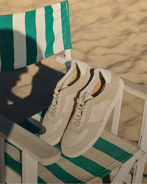 A pair of light beige suede sneakers rests on the seat of a green and white striped beach chair, with sand in the background.