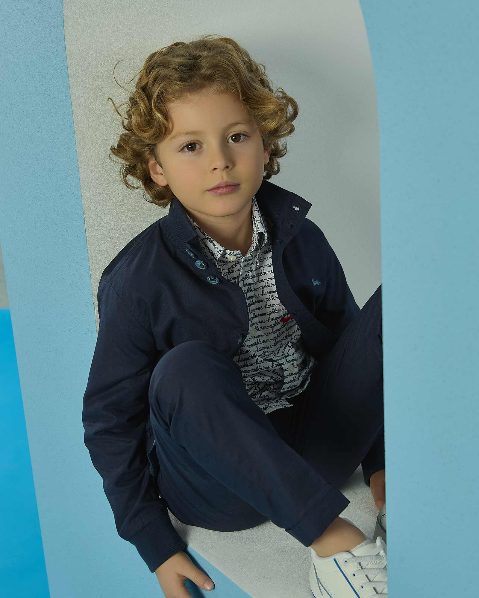 Bambino con capelli biondi in outfit blu navy seduto in una cornice azzurra.