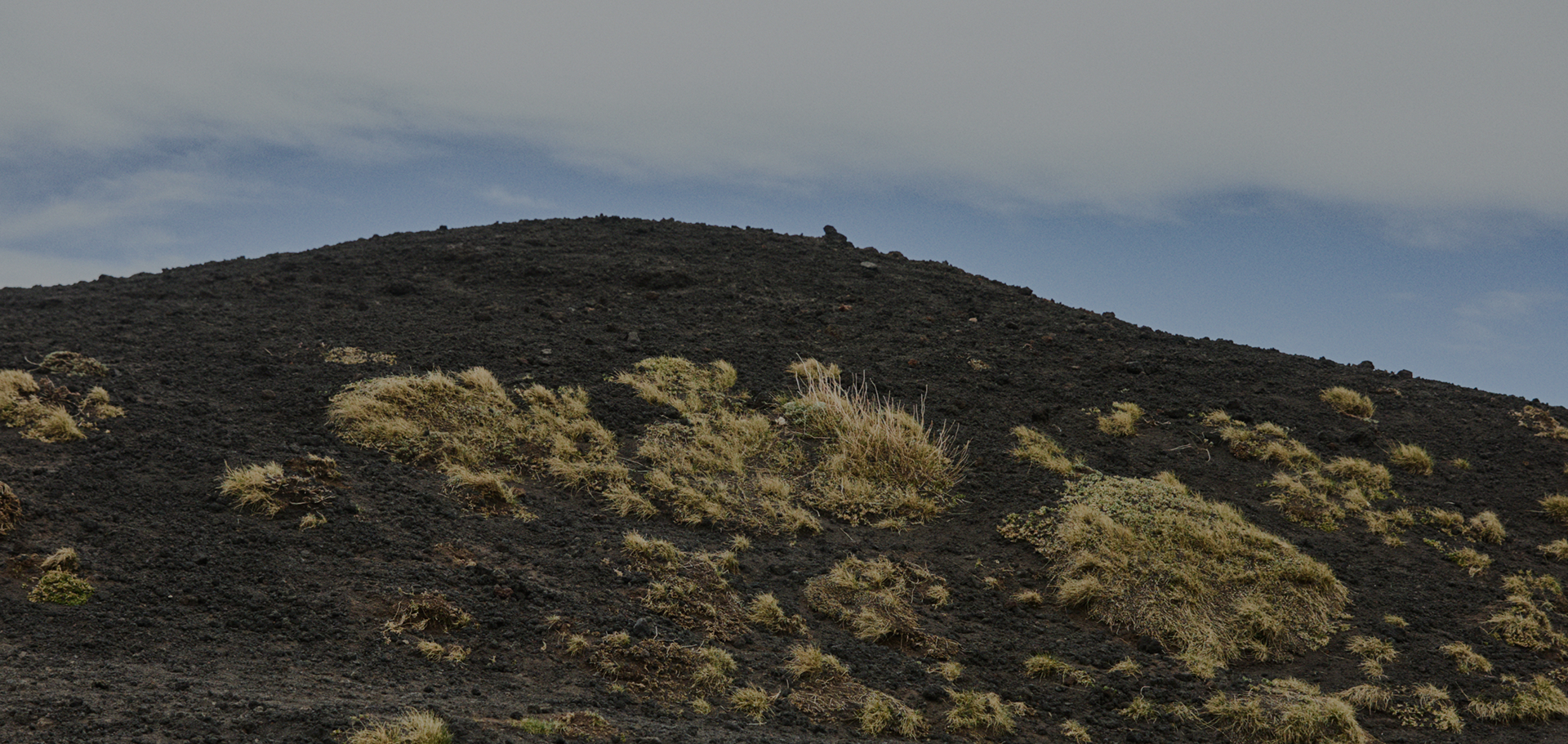 A volcanic landscape featuring a hill of dark volcanic sand and rock with small patches of dry grass, under a cloudy sky.