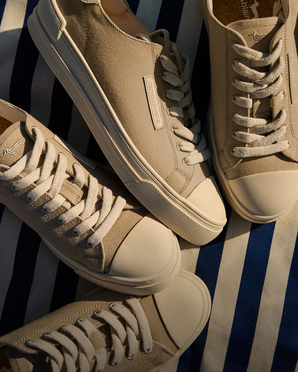 Three beige canvas sneakers with white rubber soles and toe caps are aligned horizontally on a blue and white striped cloth. Each sneaker features white laces. One of the shoes has a rectangular patch on the tongue with the "harmont & blaine" logo.