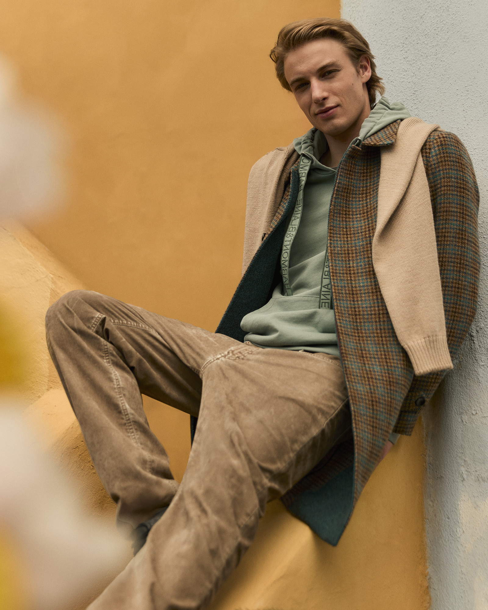 Uomo con felpa verde salvia con cappuccio e logo, cappotto a quadri e pantaloni beige