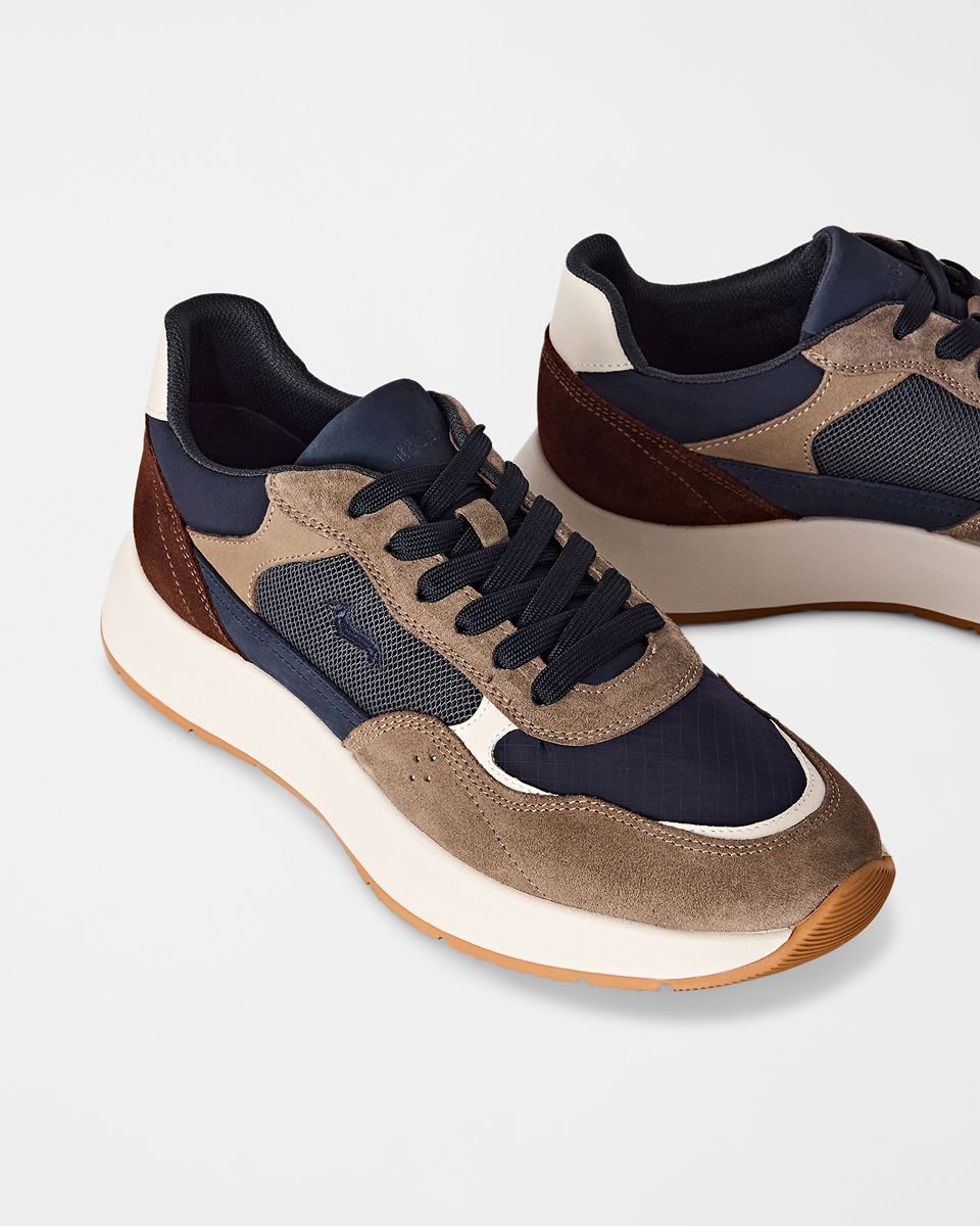 A close-up shot on a light background showing a pair of men's multi-material sneakers in navy blue, brown, and beige tones, featuring suede panels, mesh details, and a thick white and gum rubber sole.
