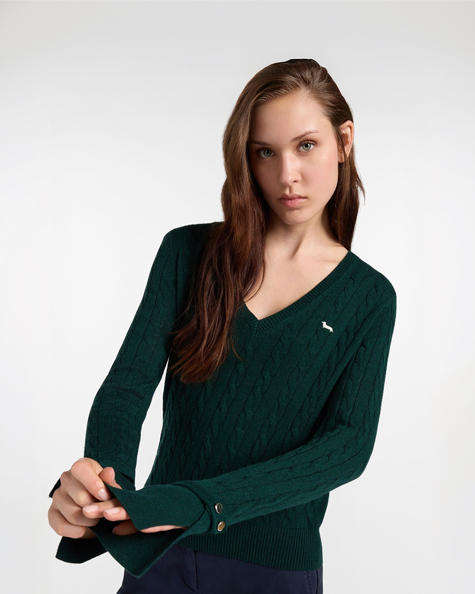 Campaign image for the Harmont & Blaine FW26 collection, focusing on women's knitwear. A model with long brown hair is featured in a half-length portrait against a neutral background, wearing a forest green cable-knit V-neck sweater. The iconic dachshund logo is embroidered in gray on the chest, while her hands in the foreground highlight the turn-back cuffs finished with two decorative gold buttons.