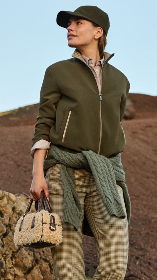 Female model wearing an olive green jacket, matching cap, patterned trousers, and a sweater tied around her waist, holding a small faux-shearling handbag against a rocky volcanic background.
