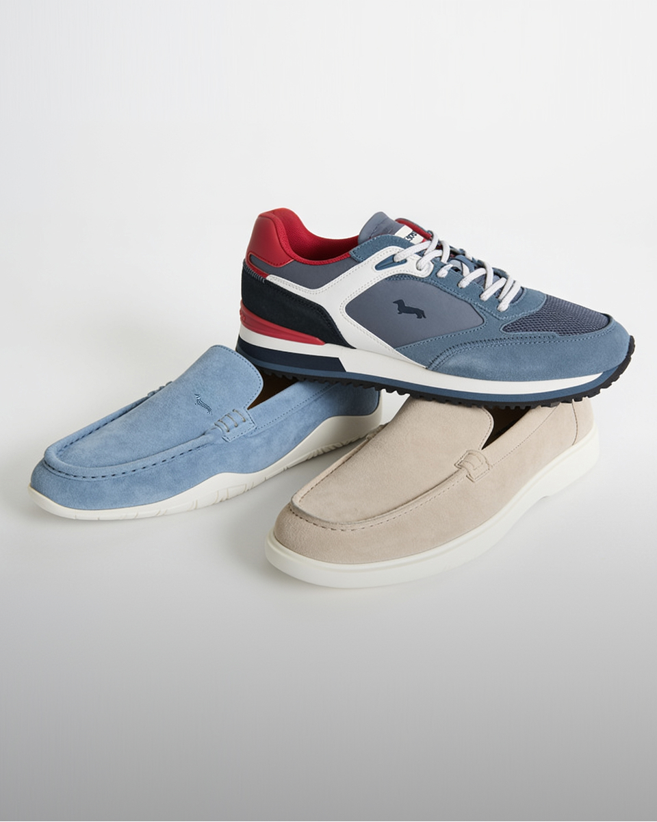 Banner Calzature Uomo: Tre modelli di scarpe disposti su un piano grigio: una sneaker tecnica blu/rossa e due mocassini scamosciati slip-on, azzurro e beige. Testo: Le Nuove Calzature. Comfort e Stile a ogni passo.