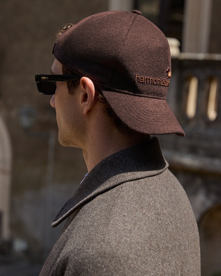 Close-up profile of a man wearing dark sunglasses and a brown wool baseball cap with an embroidered logo, paired with a grey wool coat.