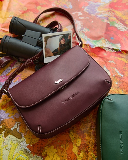 Burgundy leather crossbody bag with a metal logo resting on a geographical map, alongside black binoculars, an instant photo, and a green bag.