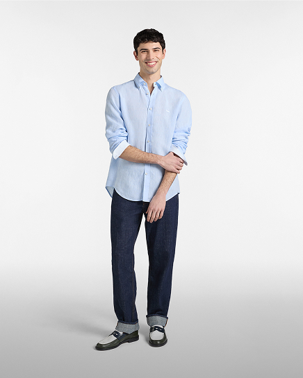 Full-length shot of a smiling male model on a white background wearing a light blue linen long-sleeved shirt with sleeves rolled up to the forearms, paired with straight-leg dark blue jeans with cuffed hems and bicolor loafers.