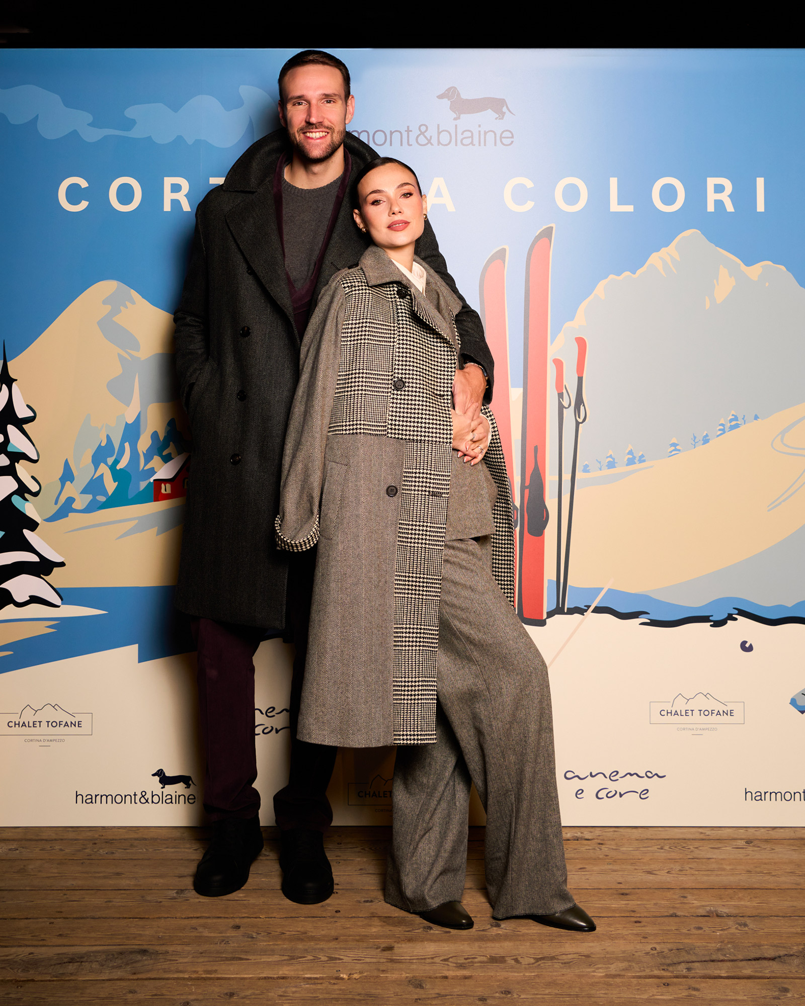 Donna in cappotto grigio a quadri e completo sartoriale con uomo in cappotto scuro davanti al backdrop Cortina a Colori.