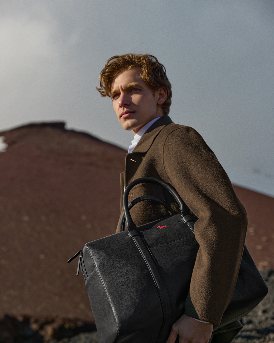 Male model wearing a brown coat and white shirt holding a large black leather travel bag under his arm, against a rocky volcanic landscape background.