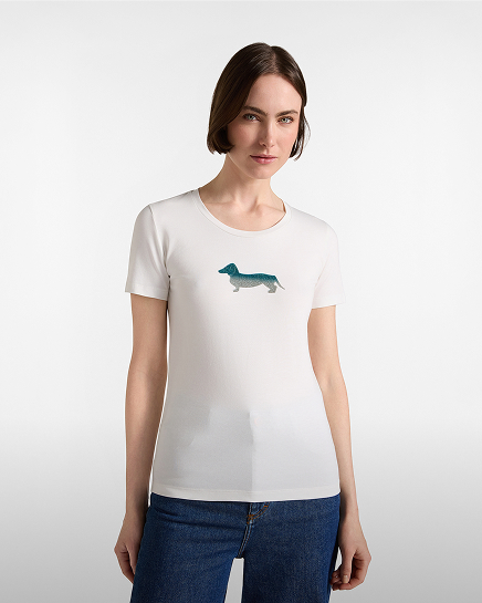 A woman with short brown hair wearing a white short-sleeved t-shirt featuring a gradient blue-to-green dachshund print on the center chest, paired with dark blue jeans.