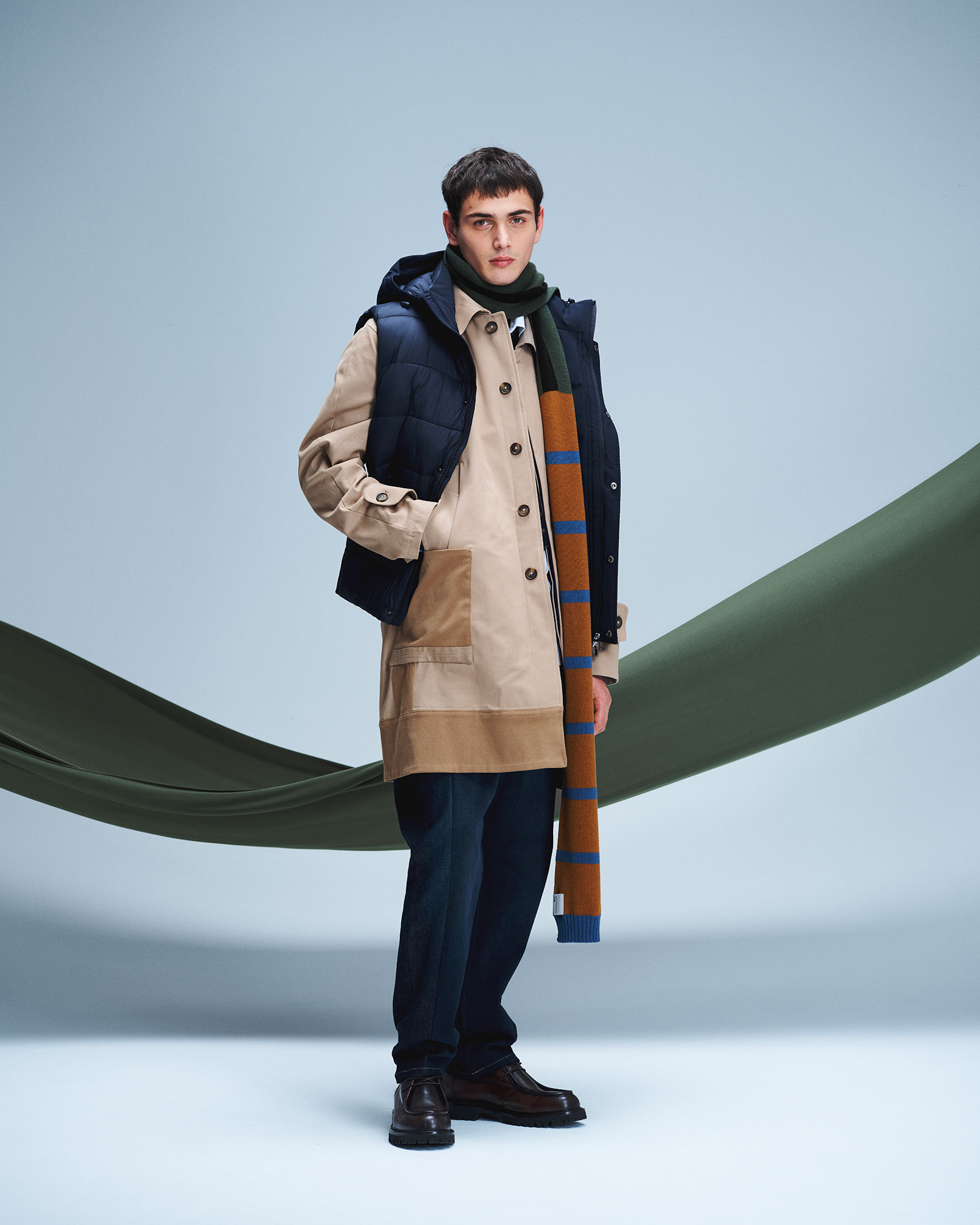 Man wearing beige parka with navy padded vest, striped scarf and dark trousers