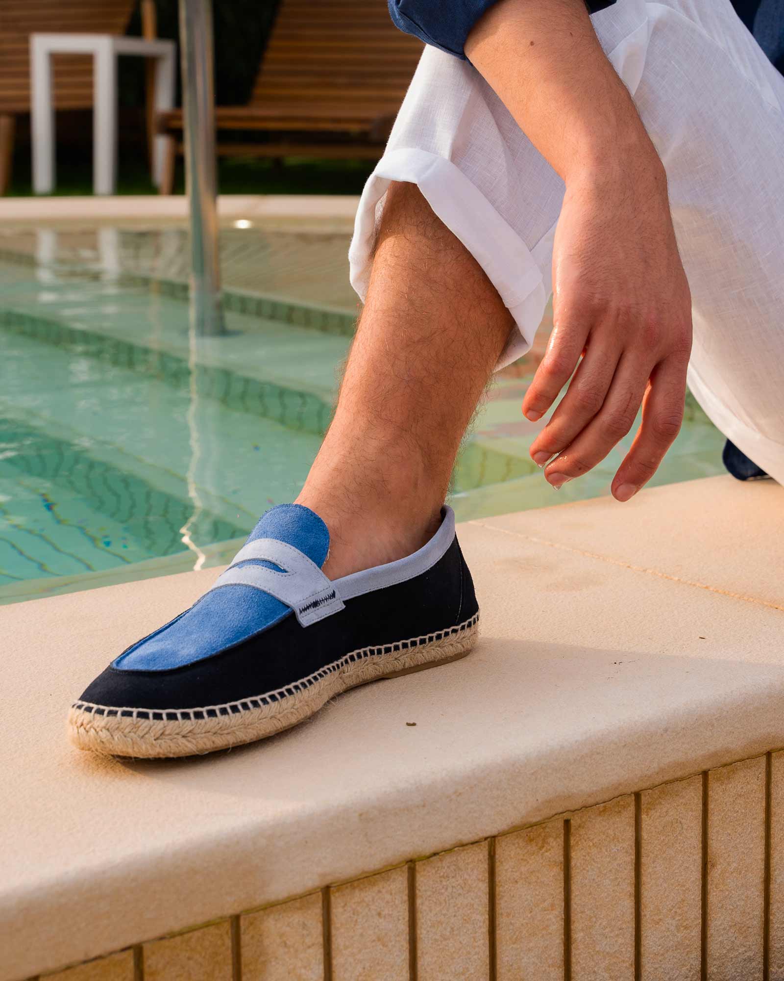 Close-up of a man's leg by the pool wearing stylish two-tone espadrilles in denim and navy blue — summer footwear elegance.