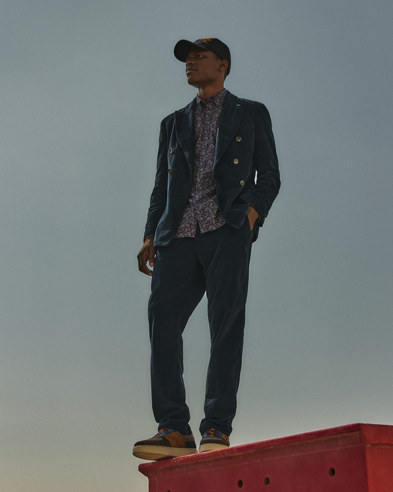 Hombre con traje de terciopelo azul y camisa estampada bajo un cielo gris.