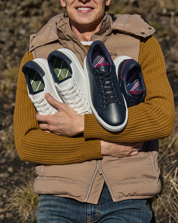 The promotional image for the Men's Footwear Focus features a smiling model holding two pairs of leather sneakers, one white pair and one navy blue pair with colorful heel accents and striped inner lining. The man wears a mustard ribbed knit sweater and a beige padded vest.