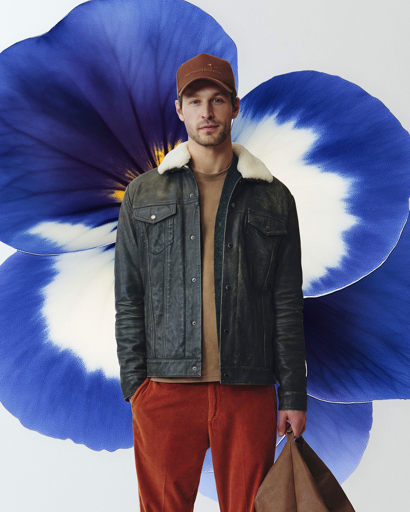 Uomo con giacca in pelle scura con collo in shearling, pantaloni in velluto a coste rossi e cappellino marrone