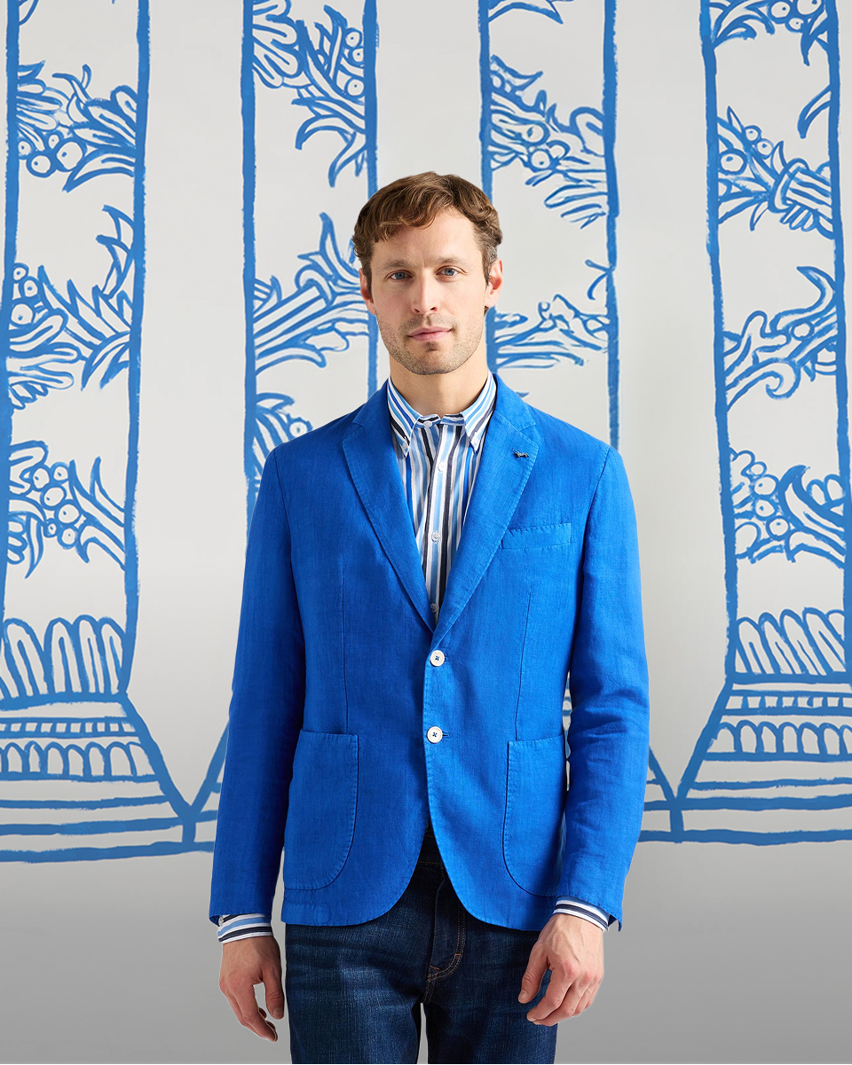 uomo in giacca di lino blu acceso, camicia a righe verticali e jeans posa davanti a uno sfondo con colonne blu disegnate. in basso compare il testo comfort d'estate e la chiamata all'azione scopri le giacche.