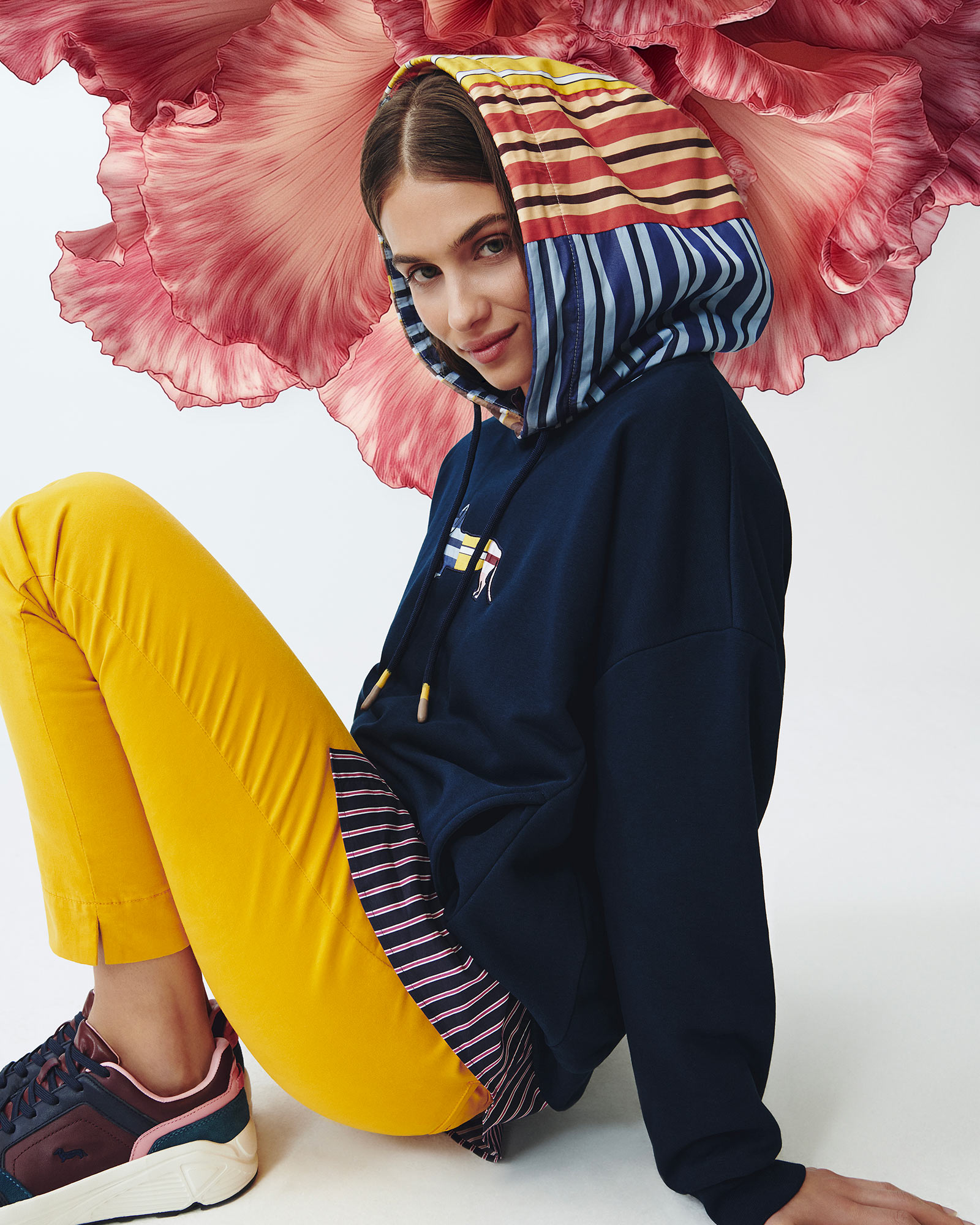 Woman wearing a dark blue hoodie with multicolored striped hood, yellow pants and multicolored sneakers.