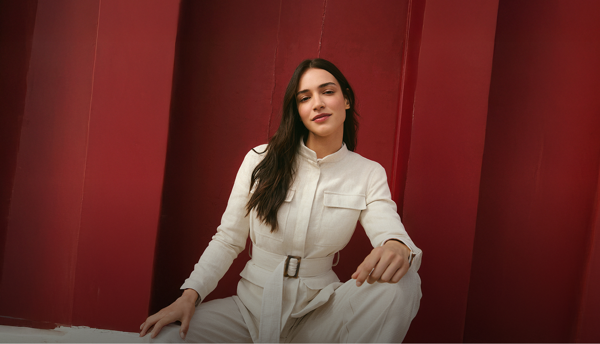 Woman in a cream-colored safari jacket with a belt, standing in front of a bright red wall. The text: made in Italy for her