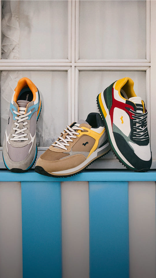 Three colorful sneakers (in grey/orange, beige/brown, and green/red/yellow tones) are displayed in a row on the light blue sill of a white window.