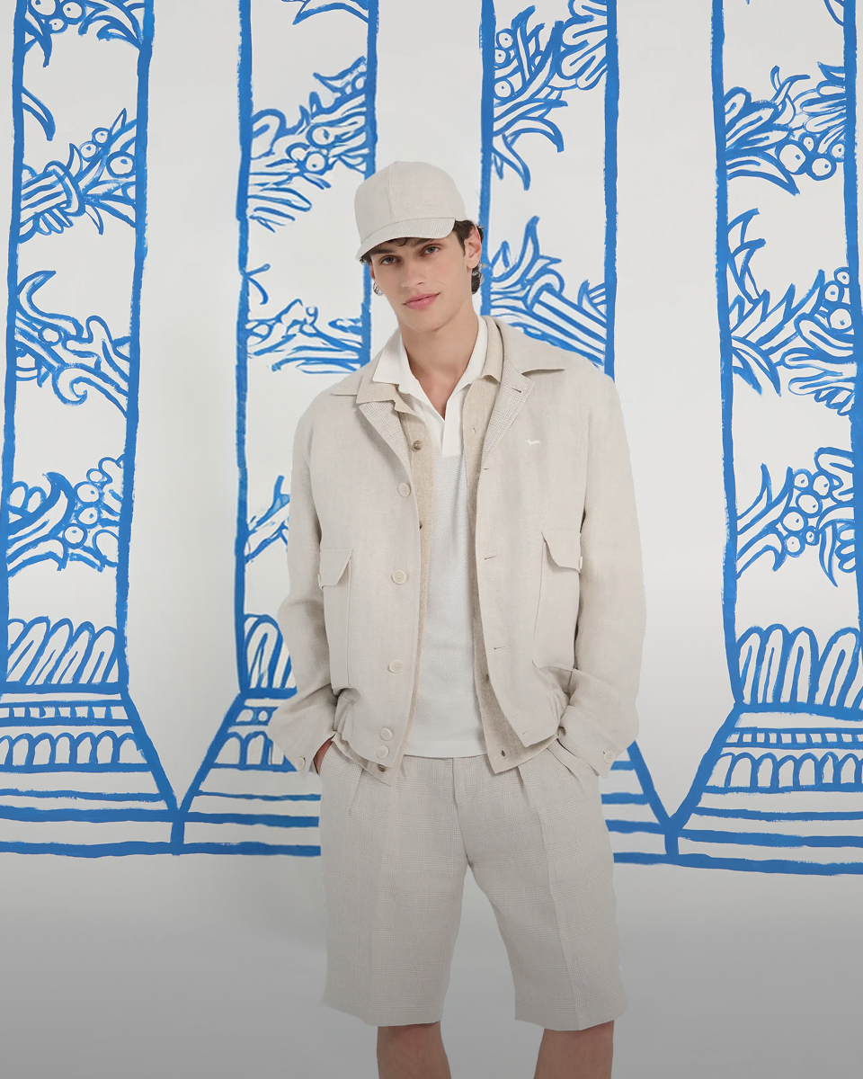 uomo in completo di lino chiaro con cappellino, giacca aperta, polo bianca e pantaloncini posa davanti a uno sfondo con colonne blu. in basso compare il testo selezione lino e la chiamata all'azione scopri ora.