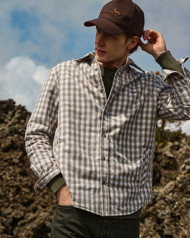 The promotional image for the Men's Shirts category features a model in a rocky outdoor setting. He wears a padded checkered overshirt in grey, beige, and light blue tones layered over an olive green sweater, paired with a brown Harmont & Blaine logo cap and dark trousers.