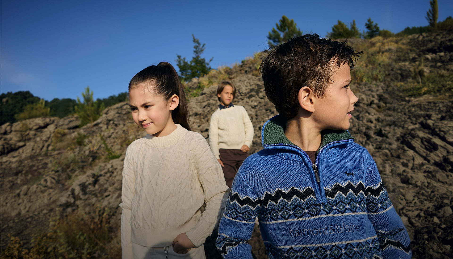 Three children are wearing Harmont&Blaine knitwear items&mdash;in the foreground, a girl in a light cable-knit sweater and a boy in a blue zip-up sweater with dark geometric patterns, while a third child in the background wears a light pullover&mdash;immersed in an outdoor natural setting under a clear blue sky.
