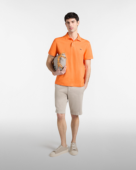Full-length shot of a male model on a bright white background wearing a vibrant orange short-sleeved polo shirt with a small embroidered dachshund logo on the chest, paired with beige chino shorts and matching low-top sneakers. The model holds a light grey crossbody bag with a decorative yellow and orange braided cord under his arm.