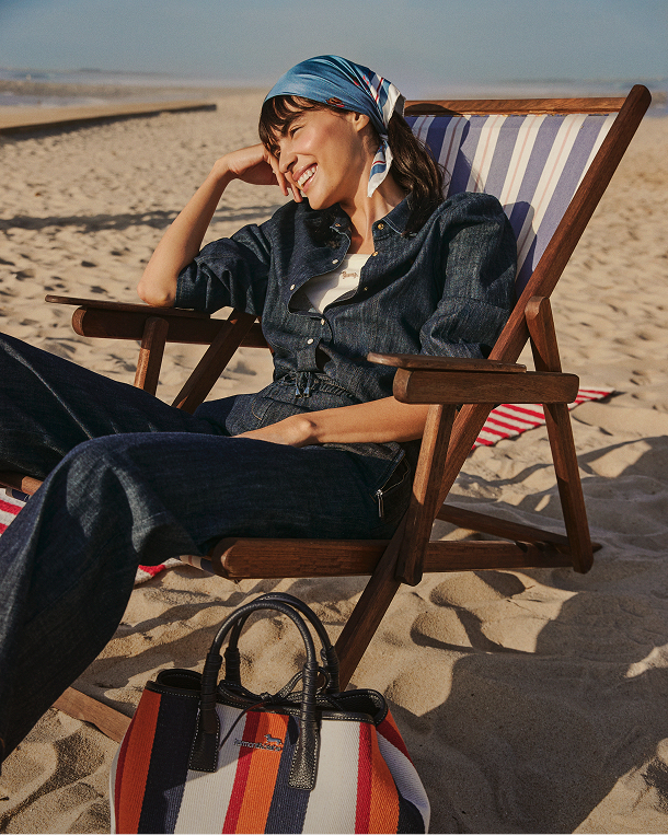 Smiling model relaxing on a deckchair at the beach, wearing a dark denim outfit, a light blue bandana, and a colored striped bag in the foreground. At the bottom, the text appears: The Weekend Essentials.