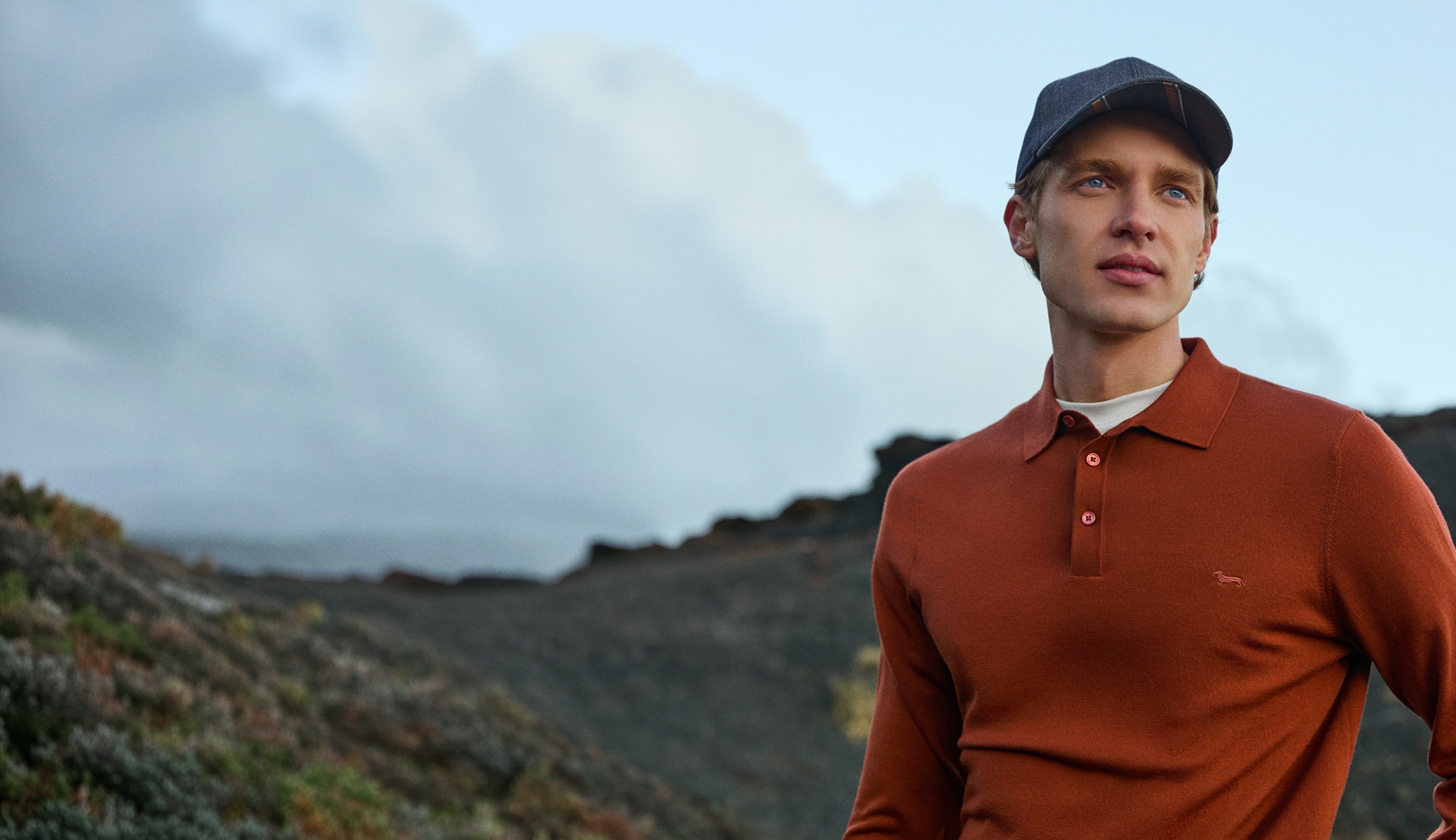 Male model wearing a rust-colored long-sleeve polo shirt and a dark baseball cap, posing outdoors in front of a hilly landscape under a cloudy sky.