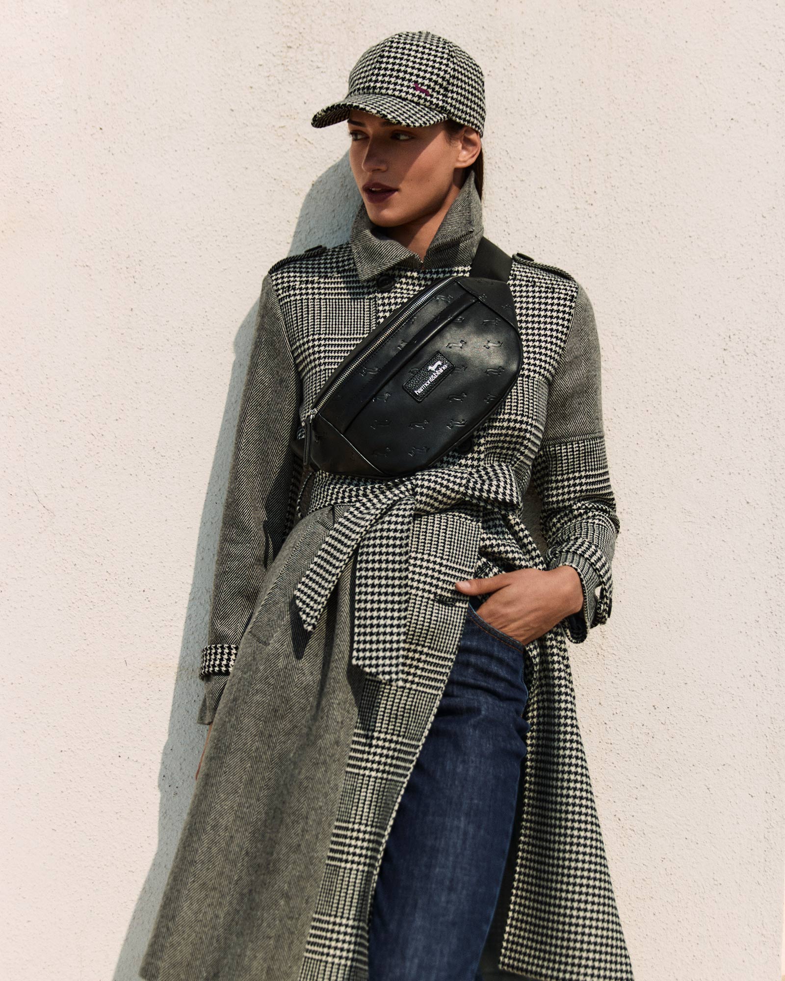 Female model wearing a houndstooth trench coat, black crossbody bag, cap and jeans.