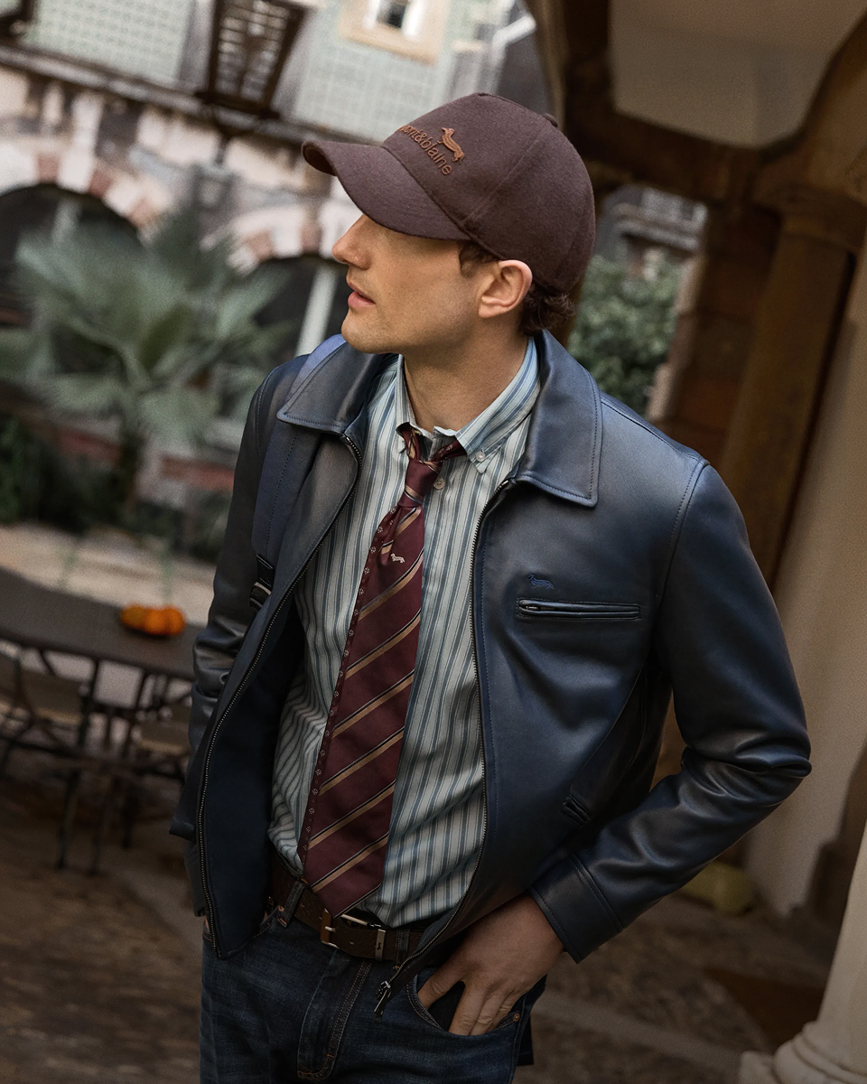 A man seen in profile wearing a dark blue leather jacket, a vertical striped shirt with a burgundy striped tie, a brown baseball cap with logo, and jeans.