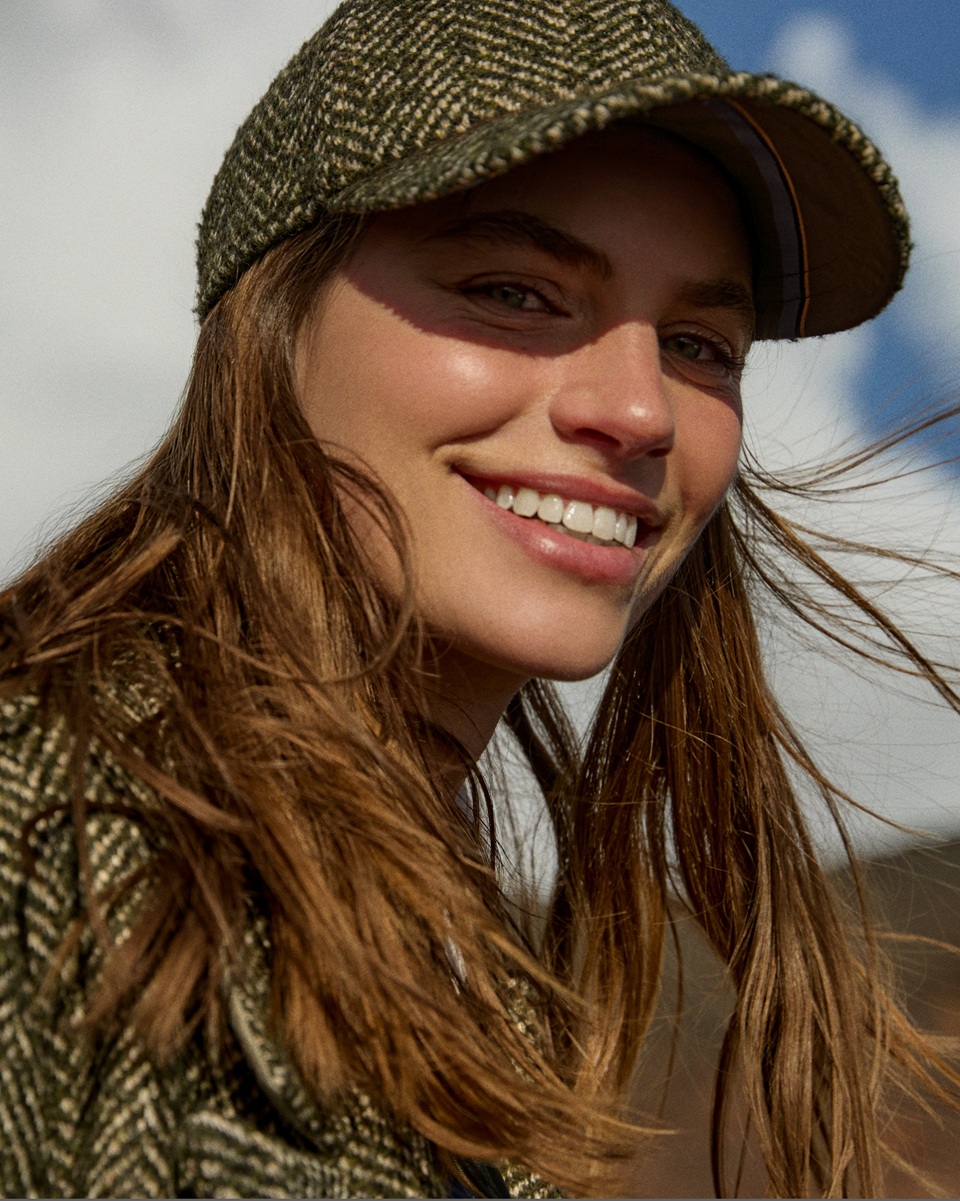 The promotional image for the Single's Day Women's campaign features a bright, close-up portrait of a model smiling toward the camera. She wears an olive green herringbone tweed baseball cap coordinating with her jacket, with long, loose hair gently blown by the breeze.
