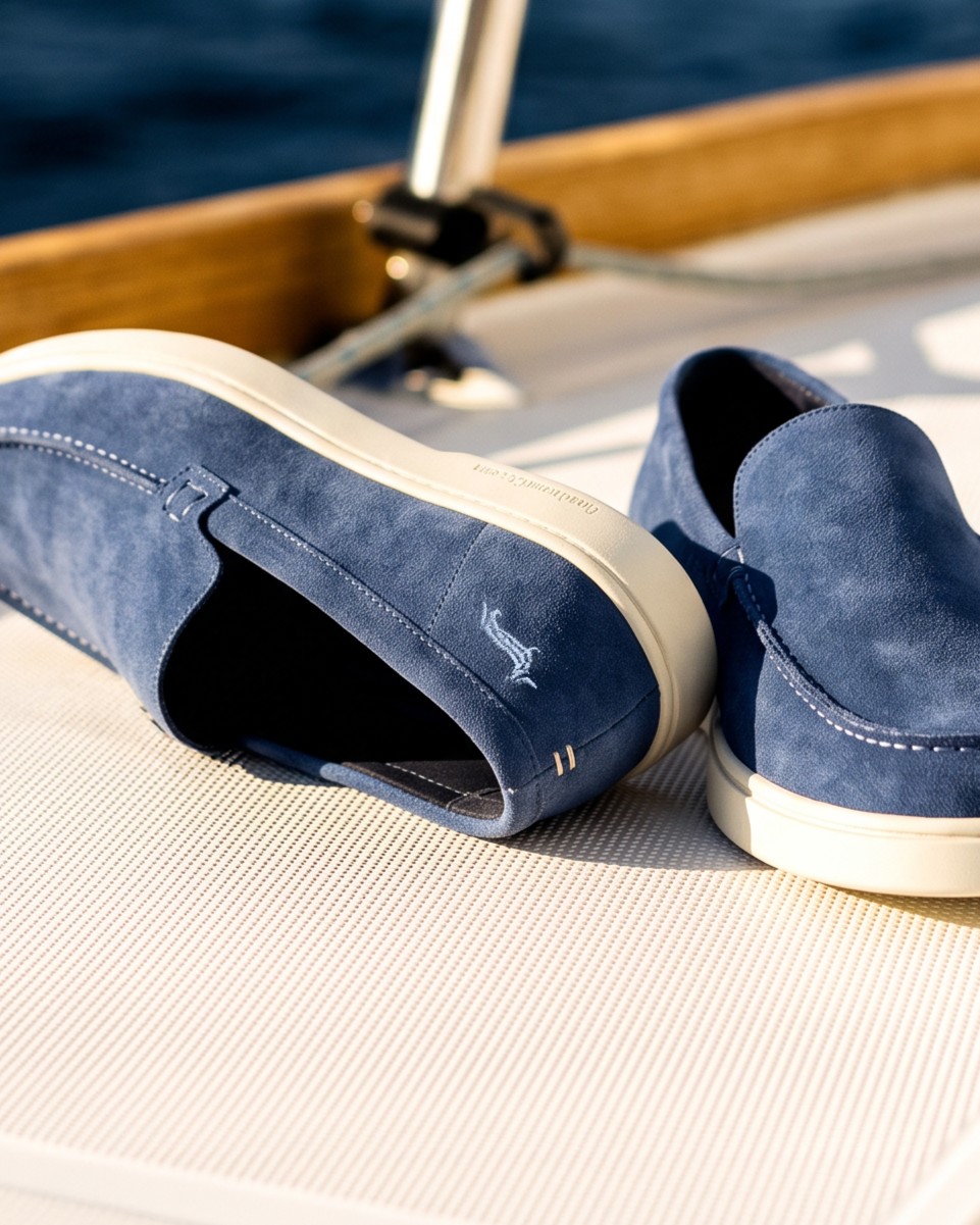 Pair of men's dark blue suede slip-on loafers featuring contrast stitching and white rubber soles, resting on the deck of a boat with the open sea and a nautical rope in the background.