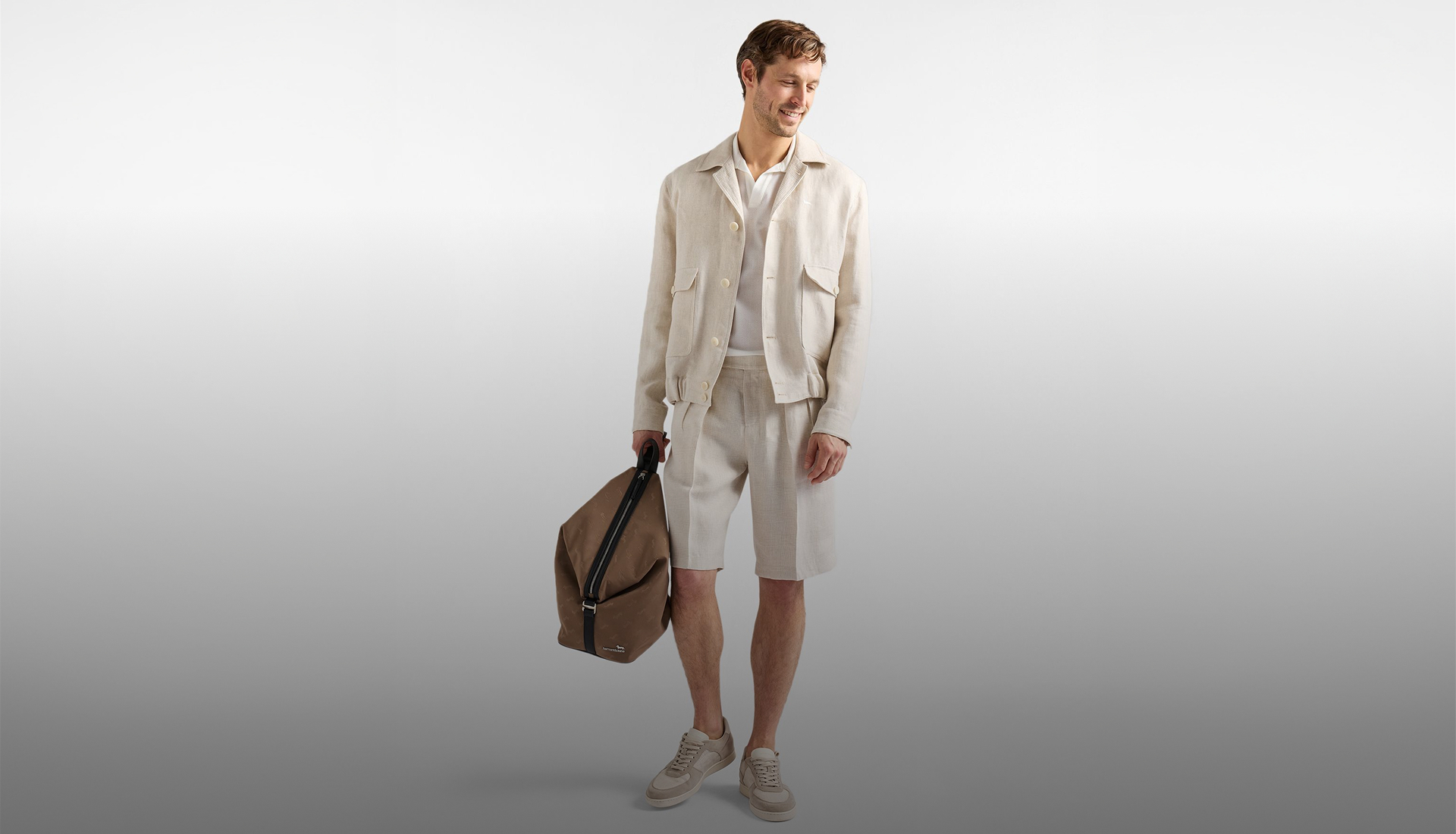 man in a light linen suit with an open jacket, white polo shirt, shorts, and sneakers looks down smiling and holds a brown bag. the background is a gradient gray. at the bottom, the text made in italy and the subtitle the authentic italian spirit appear.