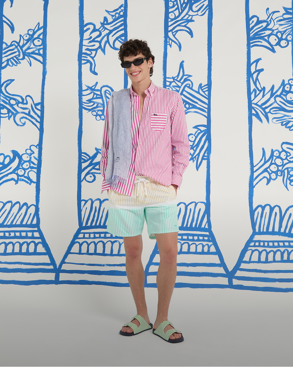 un uomo in look estivo a righe con camicia rosa e pantaloncini colorati posa davanti a uno sfondo con colonne blu. in basso compare il testo stile da spiaggia e la chiamata all'azione scopri tutti i modelli.