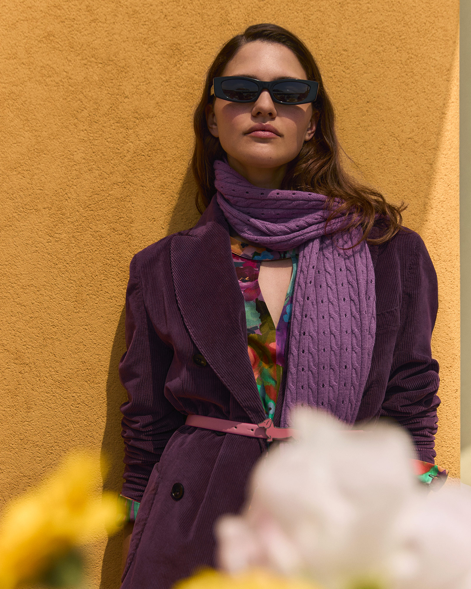 Woman wearing a purple velvet blazer, matching scarf, and black sunglasses against a yellow wall.