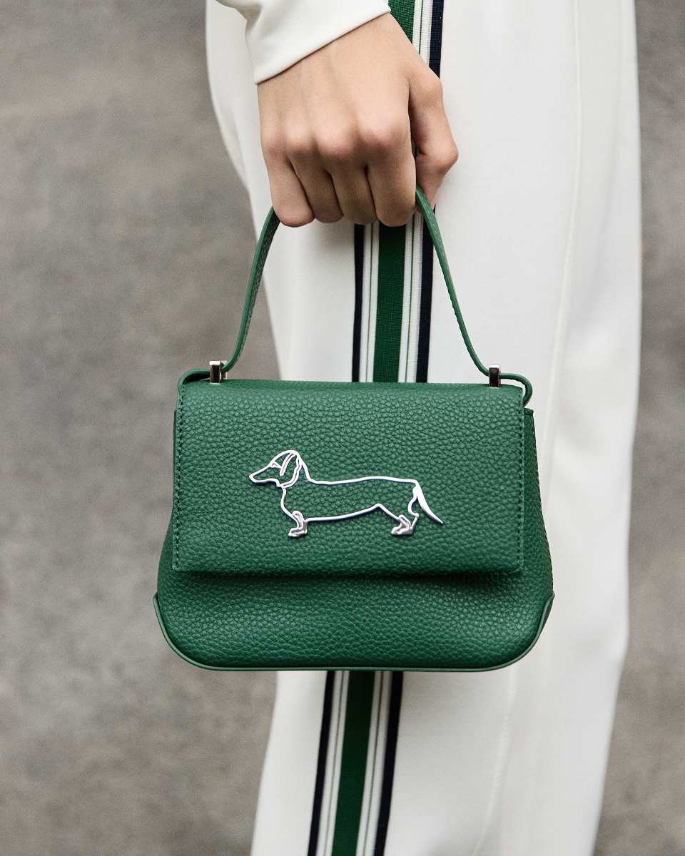 Close-up detail of a hand holding by its top handle a dark green pebbled leather handbag featuring a front flap with a metallic silver dachshund logo, paired with white trousers with green side stripes.
