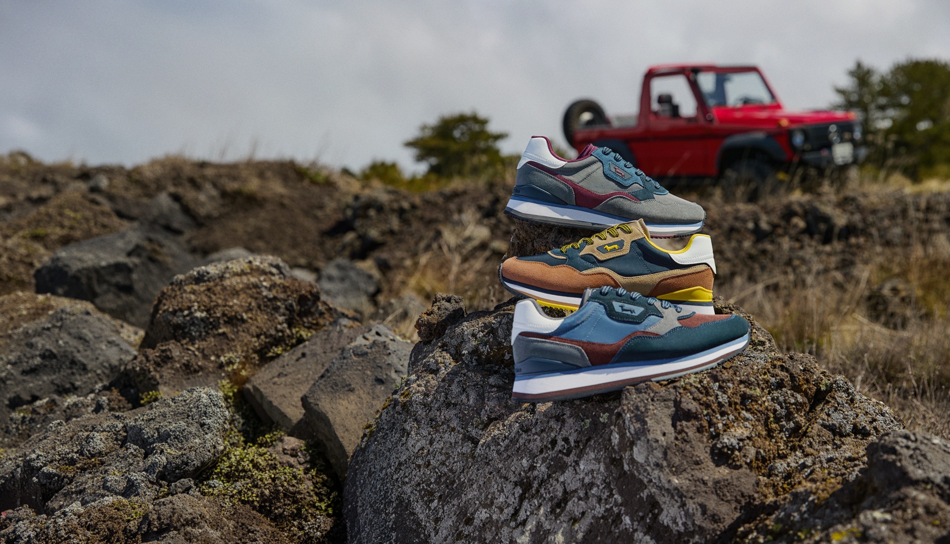 Three pairs of multicolored sneakers stacked on top of a rock, with a blurred red off-road vehicle in the background within a natural landscape.