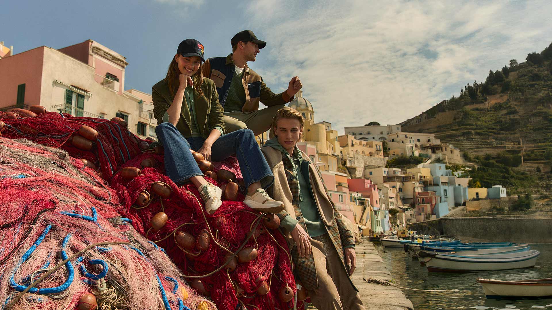 Three young models sitting on colorful fishing nets in a picturesque Mediterranean village, wearing casual layered outfits with a relaxed, coastal vibe.