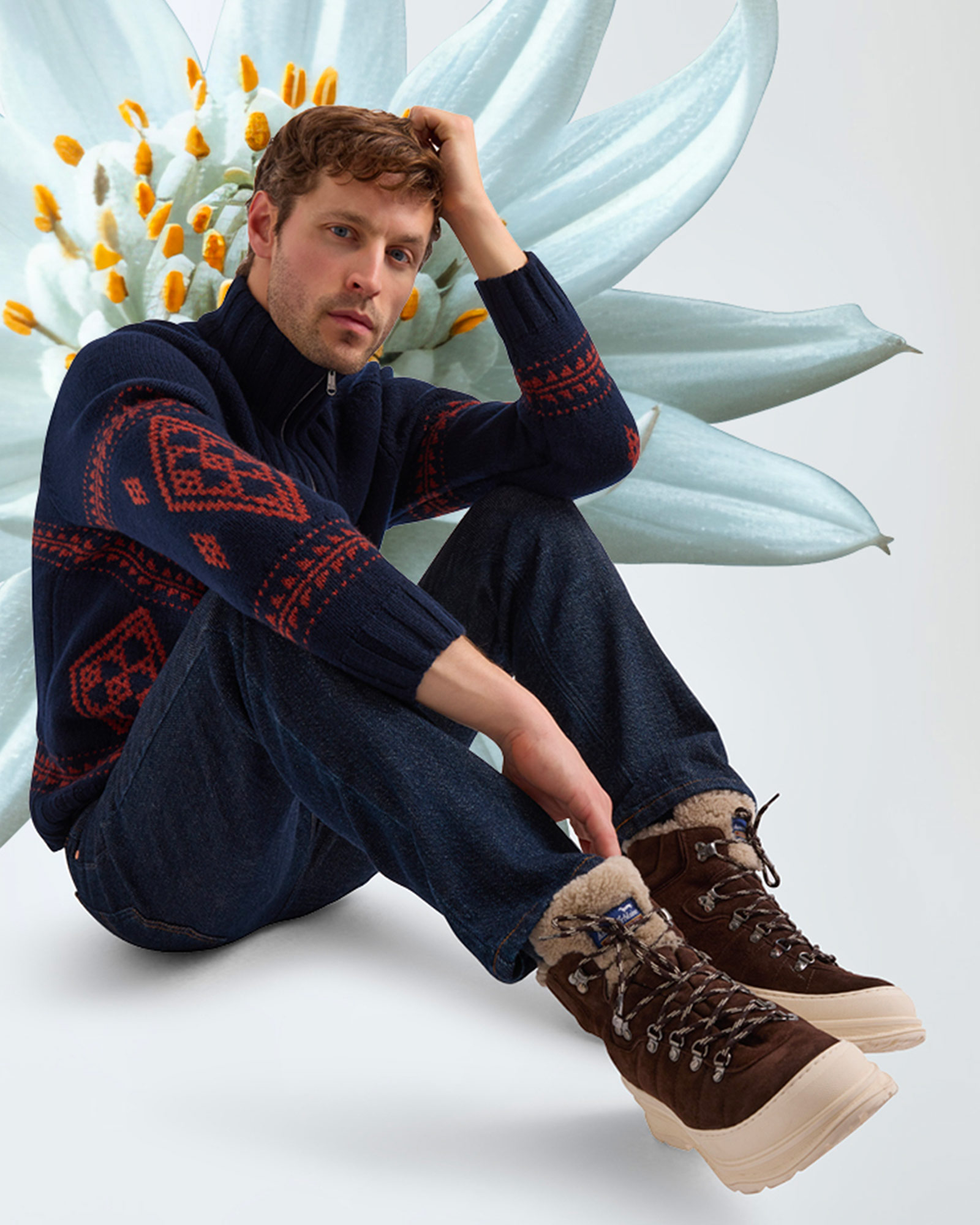 Man sitting wearing navy sweater with red patterns, dark jeans and brown boots.