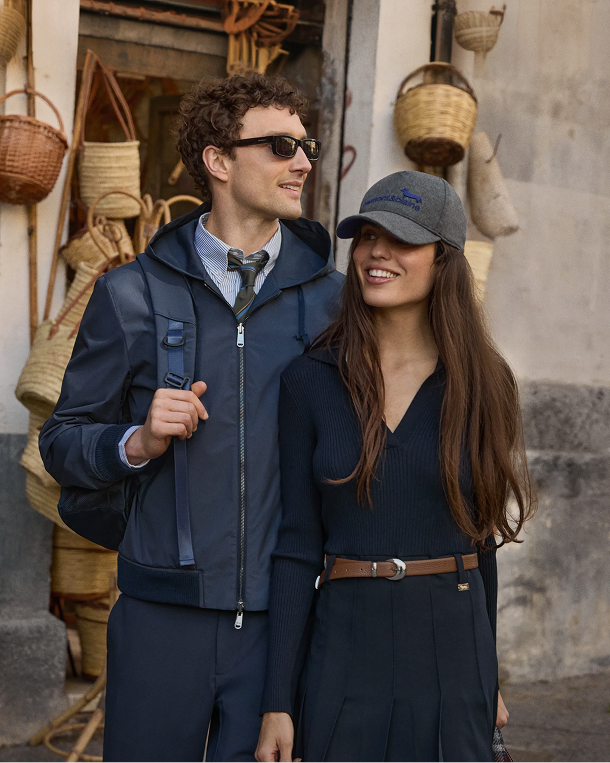 Campaign image for the Harmont & Blaine Fall/Winter 2026 preview, focusing on "Reinventing Sport". A young couple walks smiling next to a wicker basket shop: the man wears a dark blue hooded windbreaker over a shirt and tie, dark trousers, and sunglasses; the woman sports a gray cap with the brand's blue logo, a dark blue sweater and pleated skirt with a brown leather belt.