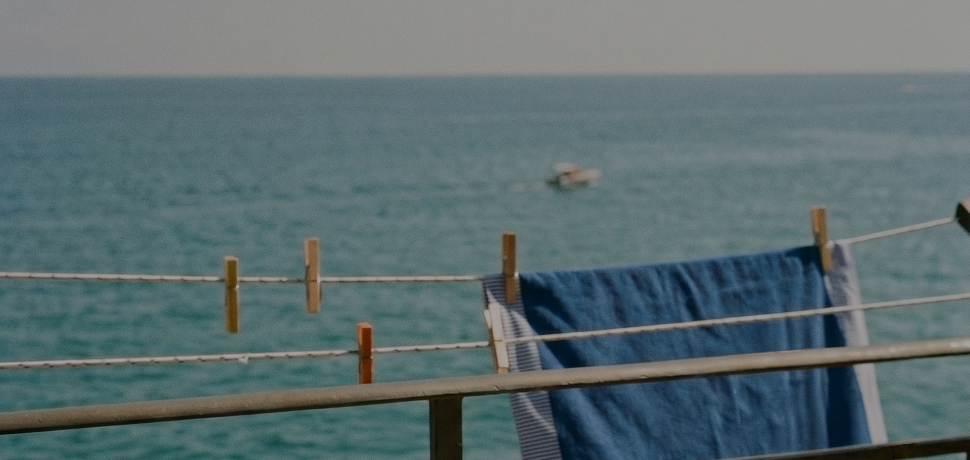 Summer view from a balcony overlooking a calm sea, with a motorboat in the distance. In the foreground, a blue beach towel with white and light blue striped borders is hanging to dry on clotheslines, held by wooden clothespins.