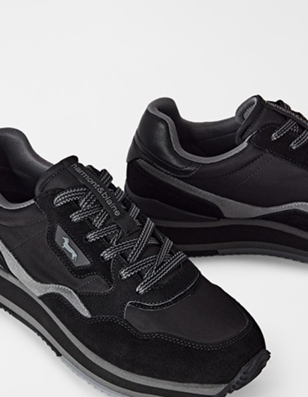 This promo image features a detailed close-up of a pair of men's sneakers in black and grey tones, crafted from a mix of technical fabrics and suede, detailed with two-tone laces and the iconic Dachshund logo on the side. The primary focus of the visual is to highlight Harmont & Blaine men's footwear, showcasing the sporty yet refined design, attention to detail, and premium material quality of the collection.
