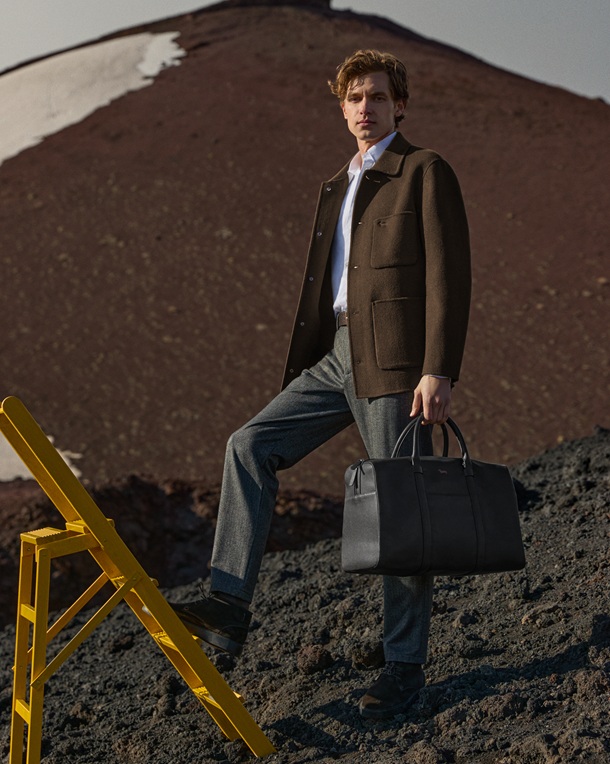 Male model wearing a brown jacket, white shirt, and grey trousers, standing on rocky volcanic ground with one foot on a yellow stepladder, holding a black travel bag.